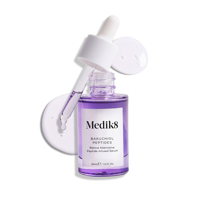 A dropper hovers above a purple glass bottle labeled "Medik8 Bakuchiol Peptides™," an age-defying serum and vitamin A alternative, surrounded by two clear liquid drops.
