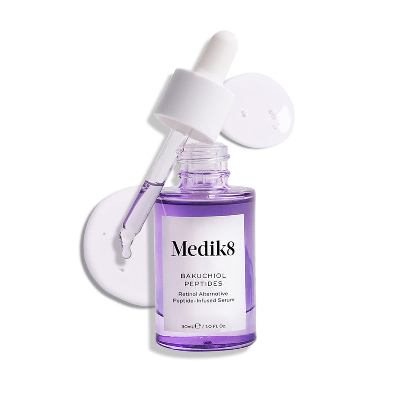 A dropper hovers above a purple glass bottle labeled "Medik8 Bakuchiol Peptides™," an age-defying serum and vitamin A alternative, surrounded by two clear liquid drops.