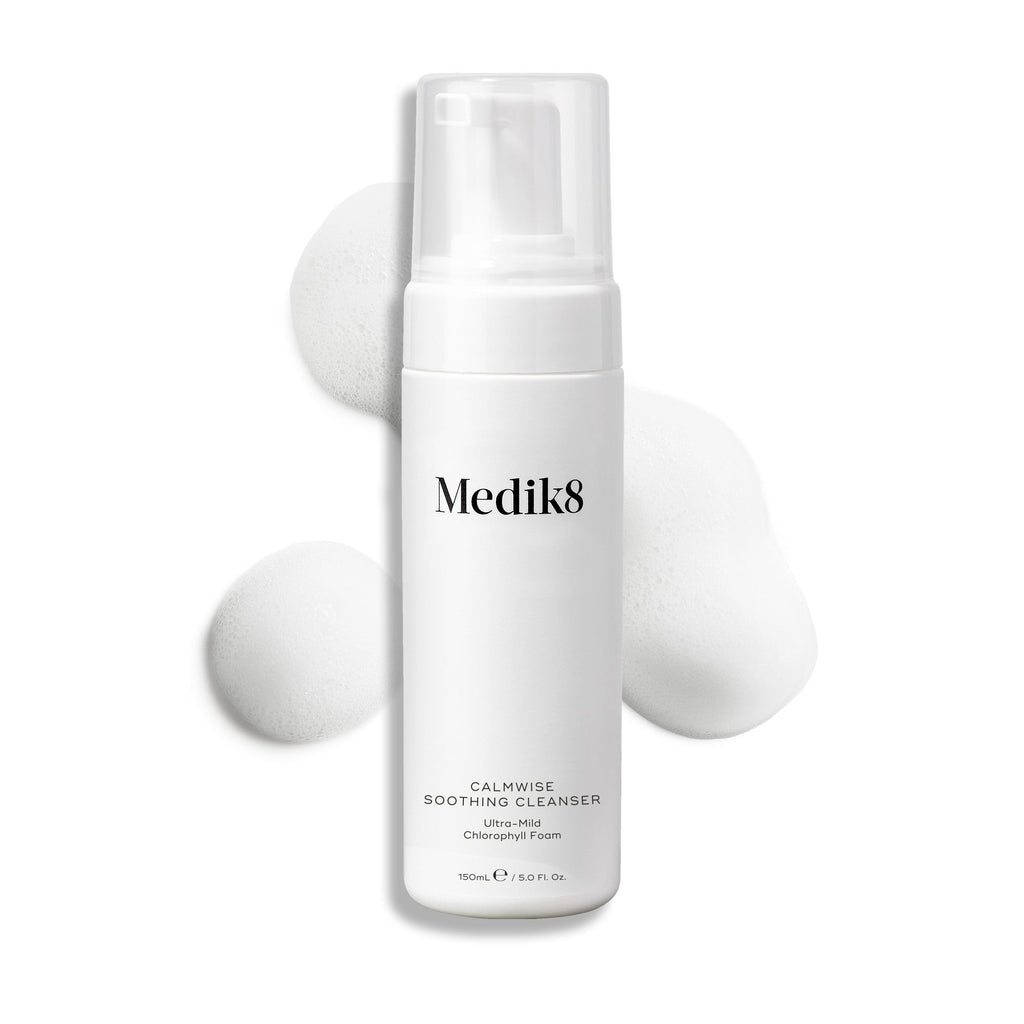 A white Medik8 Calmwise™ Soothing Cleanser bottle, chlorophyll-infused for enhanced care, features a pump top. Nestled amid gentle foam on a pristine white backdrop, it's perfect for soothing compromised skin.