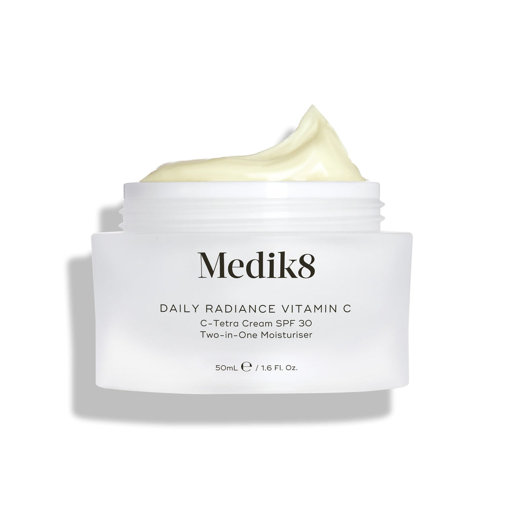A jar of Medik8 Daily Radiance Vitamin C™ cream with an open lid shows its light, creamy texture. The label promotes it as a dual-purpose moisturizer providing antioxidant protection and SPF 30 for vibrant, shielded skin.