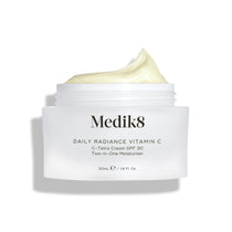 A jar of Medik8 Daily Radiance Vitamin C™ cream with an open lid shows its light, creamy texture. The label promotes it as a dual-purpose moisturizer providing antioxidant protection and SPF 30 for vibrant, shielded skin.