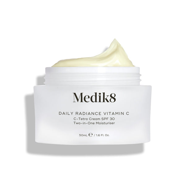 A jar of Medik8 Daily Radiance Vitamin C™ cream with an open lid shows its light, creamy texture. The label promotes it as a dual-purpose moisturizer providing antioxidant protection and SPF 30 for vibrant, shielded skin.