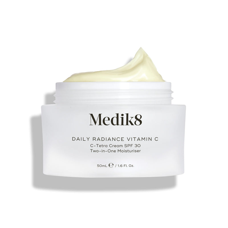 A jar of Medik8 Daily Radiance Vitamin C™ cream with an open lid shows its light, creamy texture. The label promotes it as a dual-purpose moisturizer providing antioxidant protection and SPF 30 for vibrant, shielded skin.