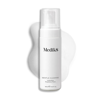 A white bottle of Medik8's Gentle Cleanse™, featuring a pump and hydrating rosemary foam, sits against a textured, foamy background. This pH-balanced cleanser is perfect for sensitive skin, providing gentle yet effective cleansing.