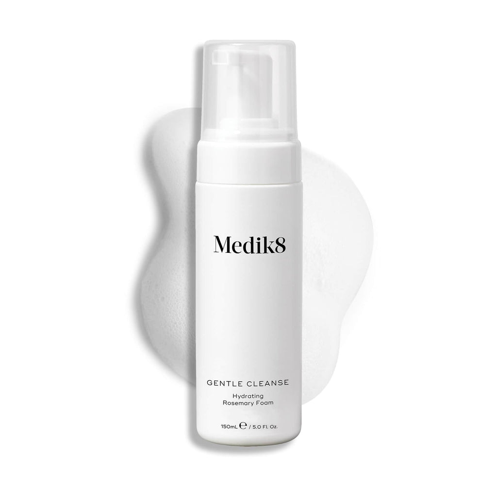 A white bottle of Medik8's Gentle Cleanse™, featuring a pump and hydrating rosemary foam, sits against a textured, foamy background. This pH-balanced cleanser is perfect for sensitive skin, providing gentle yet effective cleansing.