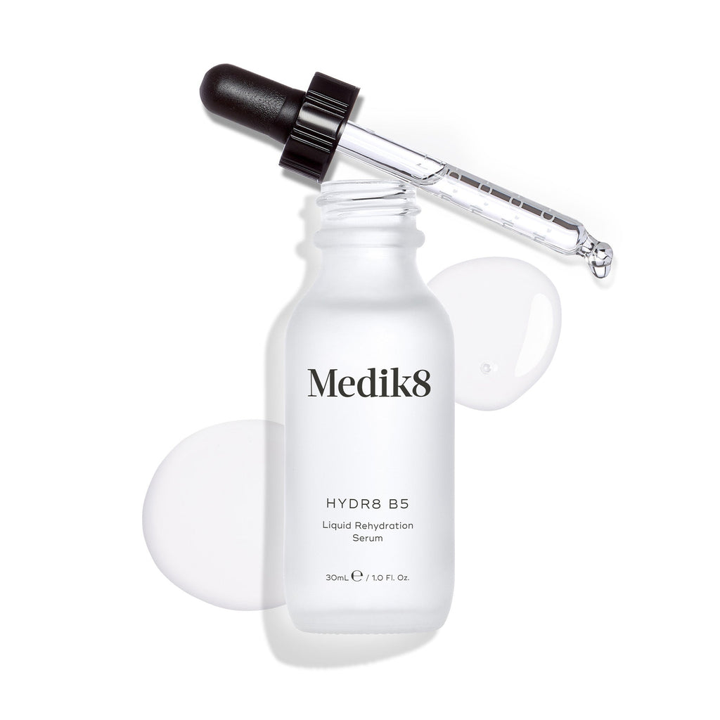 A white bottle of Medik8's Hydr8 B5™ serum, enriched with hyaluronic acid for better skin hydration, is topped with a dropper. A small amount of this fine line-reducing serum is displayed next to the bottle.