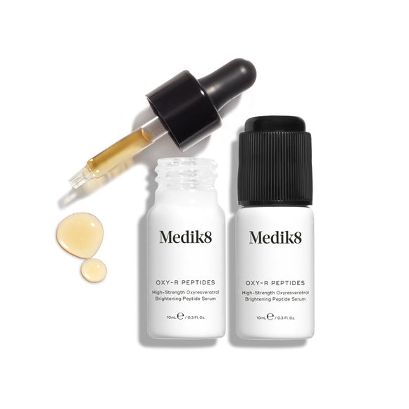 Two white Medik8 Oxy-R Peptides™ bottles with black dropper caps stand on a white background, while a dropper releases oxyresveratrol-infused serum designed to target dark spots and rejuvenate skin with peptide power.-9