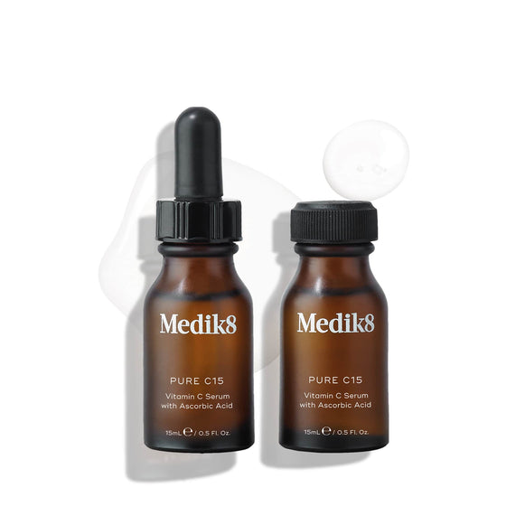 Two brown bottles of Medik8 Pure C15™ featuring a black dropper rest on a white background. A small drop of the age-defying serum, known for its antioxidant protection with ascorbic acid, is visible beside them.-7