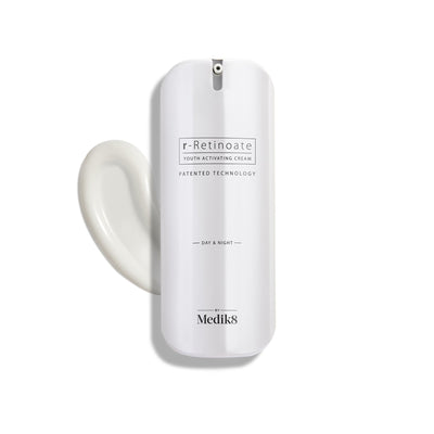 A white bottle of Medik8 r-Retinoate® Day & Night cream, containing retinyl retinoate and vitamin A, comes with a pump dispenser and is pictured next to a swirl of the anti-aging serum.