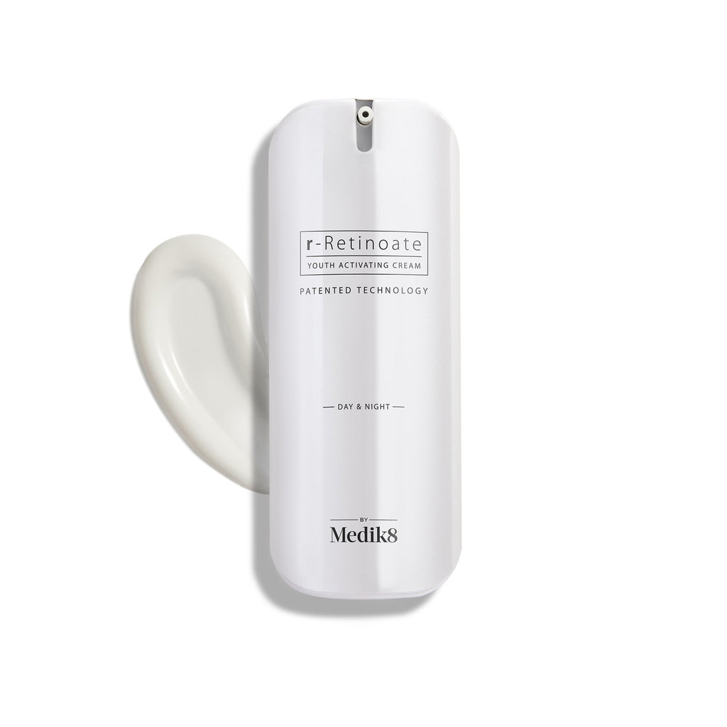 A white bottle of Medik8 r-Retinoate® Day & Night cream, containing retinyl retinoate and vitamin A, comes with a pump dispenser and is pictured next to a swirl of the anti-aging serum.
