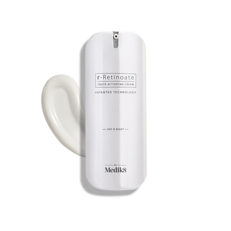 A white bottle of Medik8 r-Retinoate® Day & Night cream, containing retinyl retinoate and vitamin A, comes with a pump dispenser and is pictured next to a swirl of the anti-aging serum.