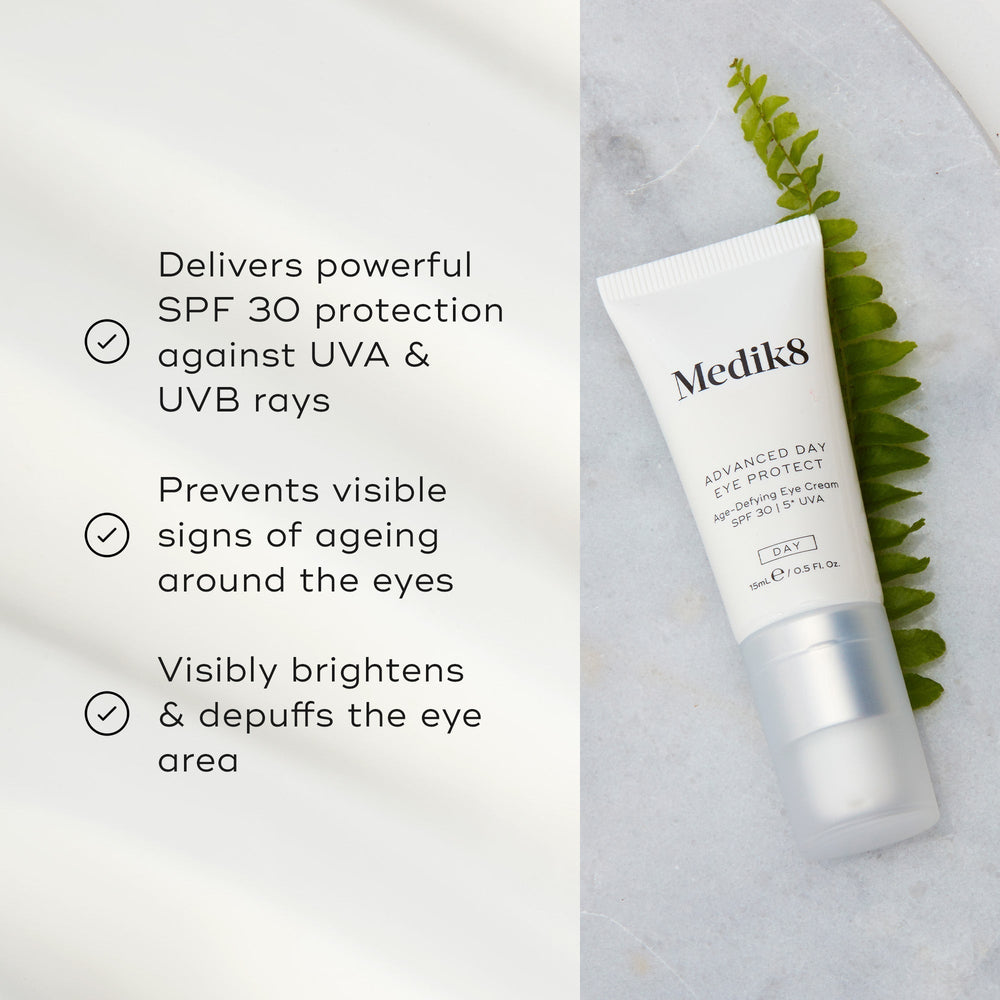 A tube of Medik8's Advanced Day Eye Protect™ cream is on a marble surface next to a leaf, highlighting its SPF 30 protection, anti-aging properties, and brightening effects for effective wrinkle prevention.