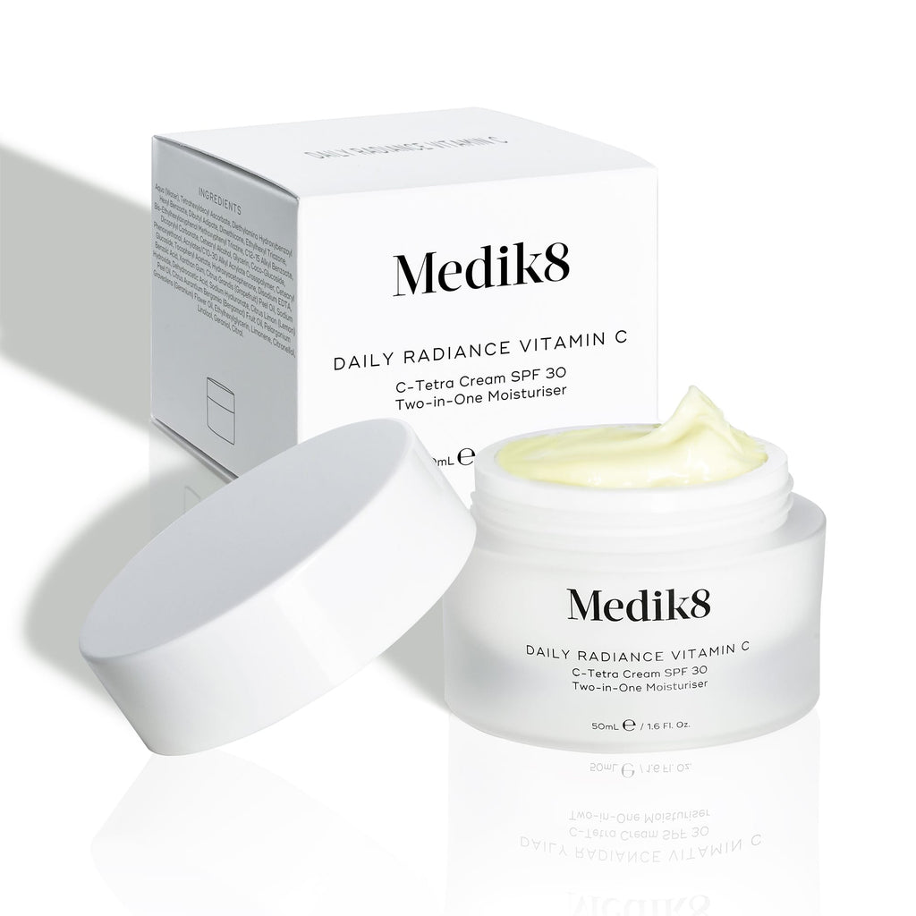 An open jar of Medik8's Daily Radiance Vitamin C™ cream reveals its light yellow contents, offering SPF 30 and antioxidant protection, with the product box in the background.