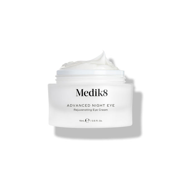 A small white jar labeled "Advanced Night Eye™" by Medik8 reveals its rejuvenating nighttime eye cream, rich in vitamin A, with the lid off and cream visible.-