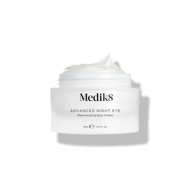 A small white jar labeled "Advanced Night Eye™" by Medik8 reveals its rejuvenating nighttime eye cream, rich in vitamin A, with the lid off and cream visible.