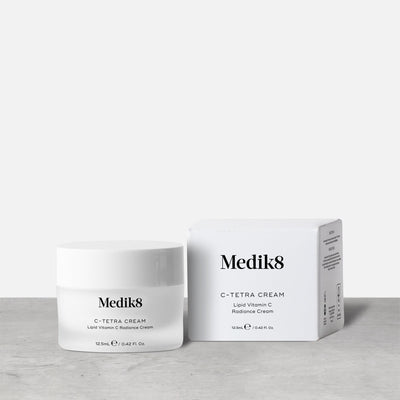 A jar of Medik8's C-Tetra® Cream, enriched with vitamin C, is placed next to its box on a gray surface against a plain white background.