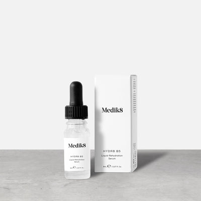 A Medik8 Hydr8 B5™ Liquid Rehydration Serum, equipped with a dropper and enriched with hyaluronic acid for superior skin hydration, is positioned next to its white packaging on a gray surface against a plain background.