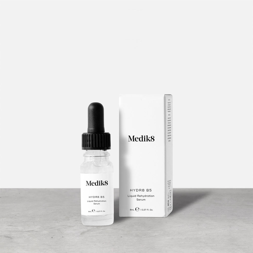 A Medik8 Hydr8 B5™ Liquid Rehydration Serum, equipped with a dropper and enriched with hyaluronic acid for superior skin hydration, is positioned next to its white packaging on a gray surface against a plain background.