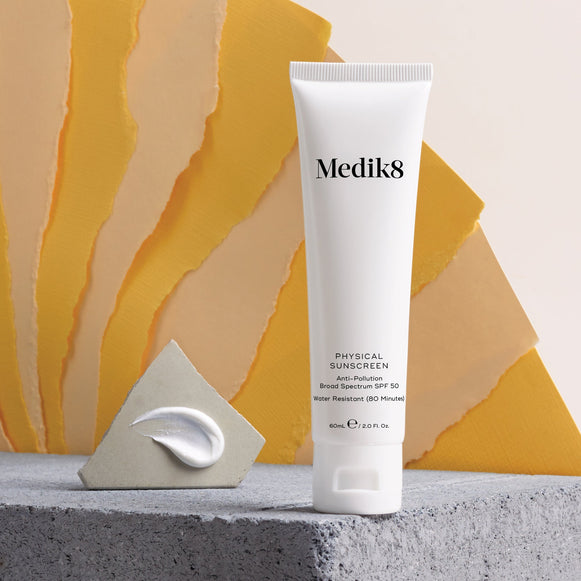 The Medik8 Physical Sunscreen™, with SPF50 UVA/UVB protection, is displayed on a concrete surface beside a small triangular block with a cream smear, set against a backdrop of yellow fan-like elements.-hover-3